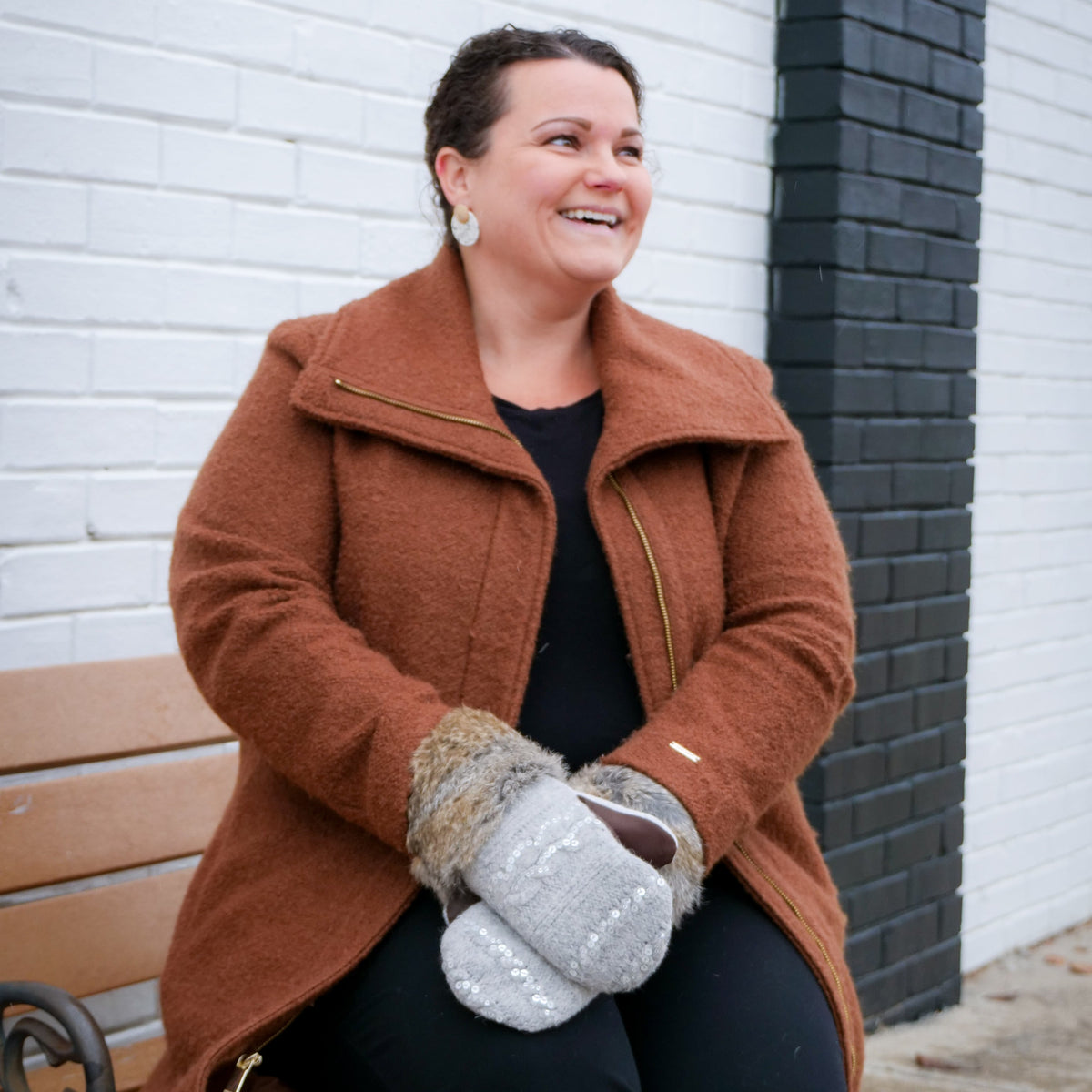 A woman sitting on a wooden bench outdoors, wearing a brown wool-like coat, black outfit, and gray knit mittens, with a white brick wall and black brick column behind her.