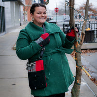 A woman wearing a green wool coat, red and gray knit gloves, and a black crossbody bag with red accents stands on a city sidewalk decorated for Christmas.