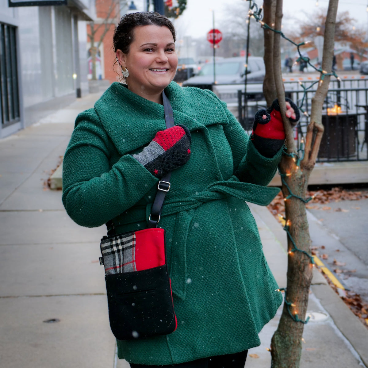 A woman wearing a green wool coat, red and gray knit gloves, and a black crossbody bag with red accents stands on a city sidewalk decorated for Christmas.