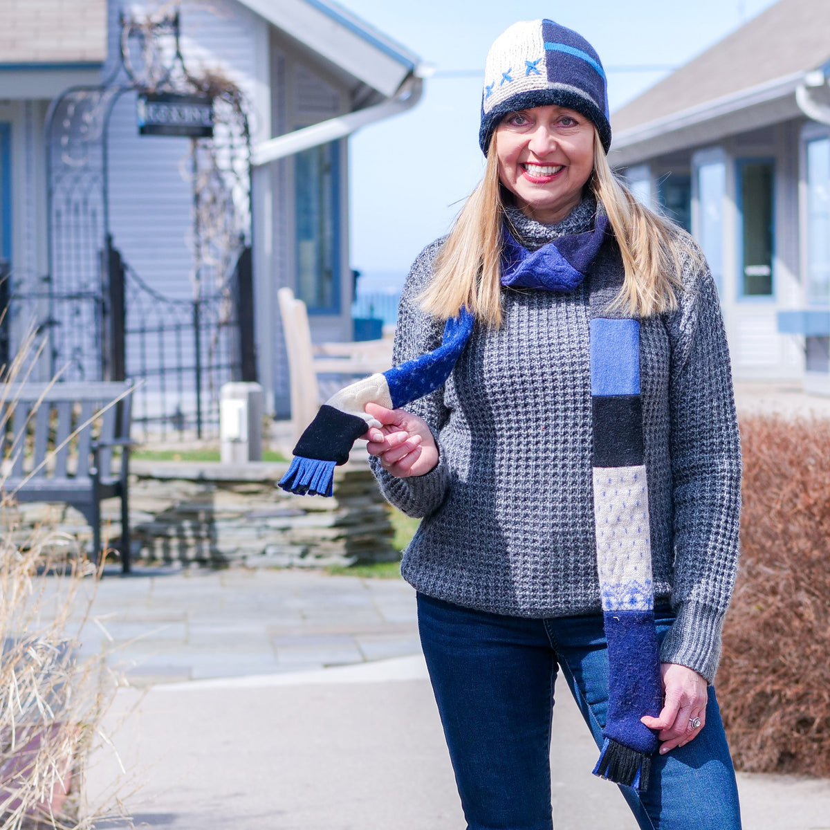 Sue Burns wearing a gray knit sweater, a blue and white baabaazuzu scarf, a matching blue and white knit beanie, and dark jeans stands outdoors in Leelanau County.