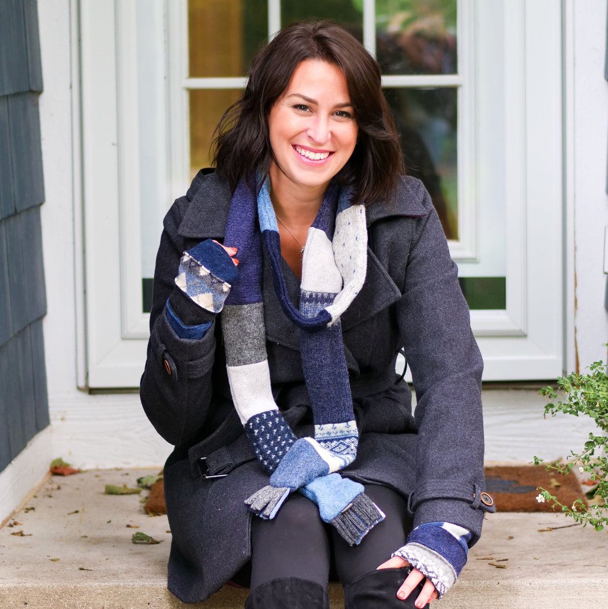 A woman sitting on concrete steps in front of a house, wearing a dark charcoal coat, a chunky blue, gray, and white skinny scarf, and black leggings.