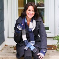 A woman sitting on concrete steps in front of a house, wearing a dark charcoal coat, a chunky blue, gray, and white skinny scarf, and black leggings.