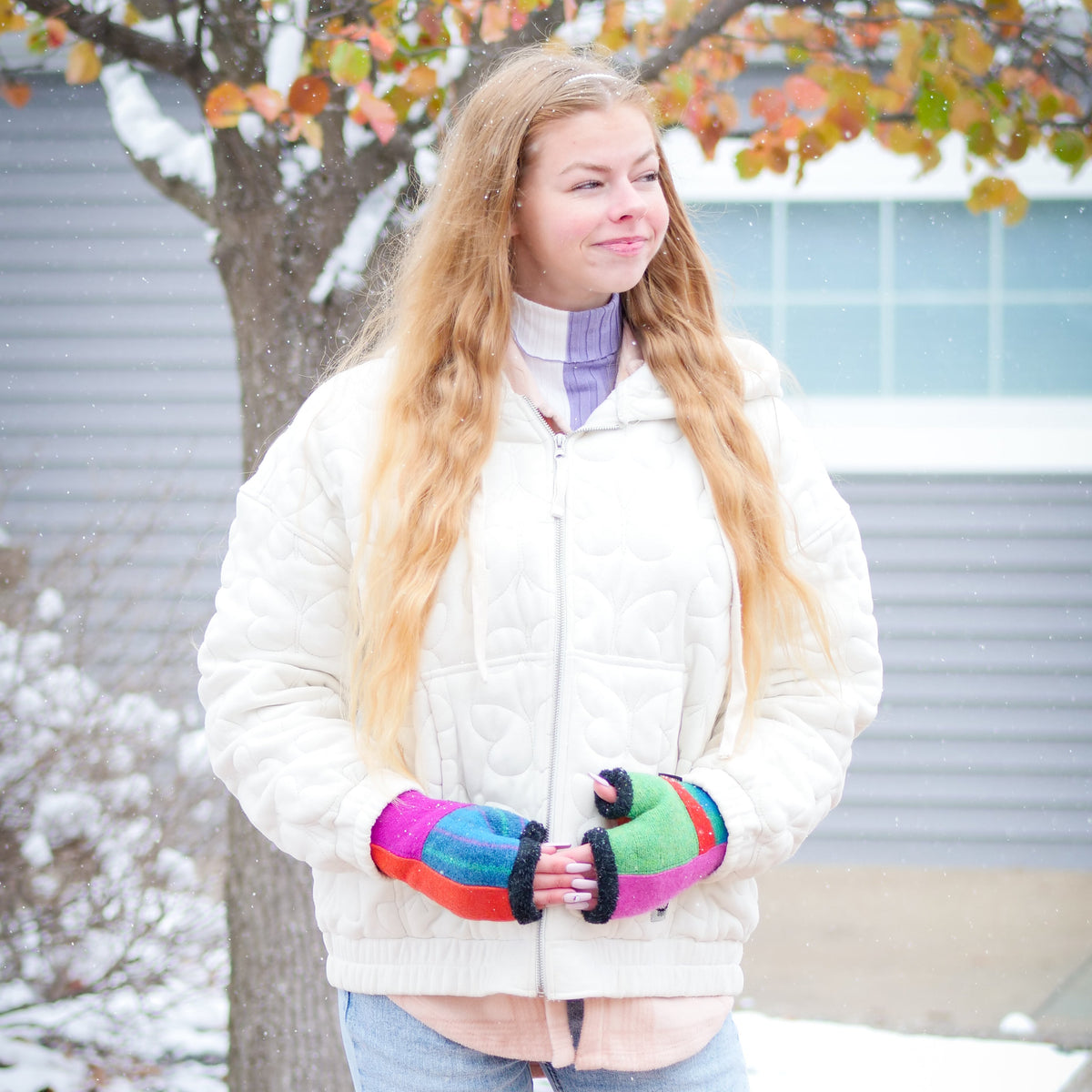 A young woman standing outdoors in a snowy setting, wearing a cream quilted jacket, light blue jeans, and a purple turtleneck underneath her jacket. She is holding a pair of vibrant, multicolored knit fingerless gloves.
