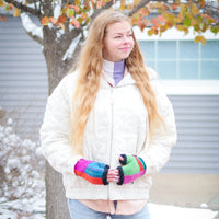 A young woman standing outdoors in a snowy setting, wearing a cream quilted jacket, light blue jeans, and a purple turtleneck underneath her jacket. She is holding a pair of vibrant, multicolored knit fingerless gloves.