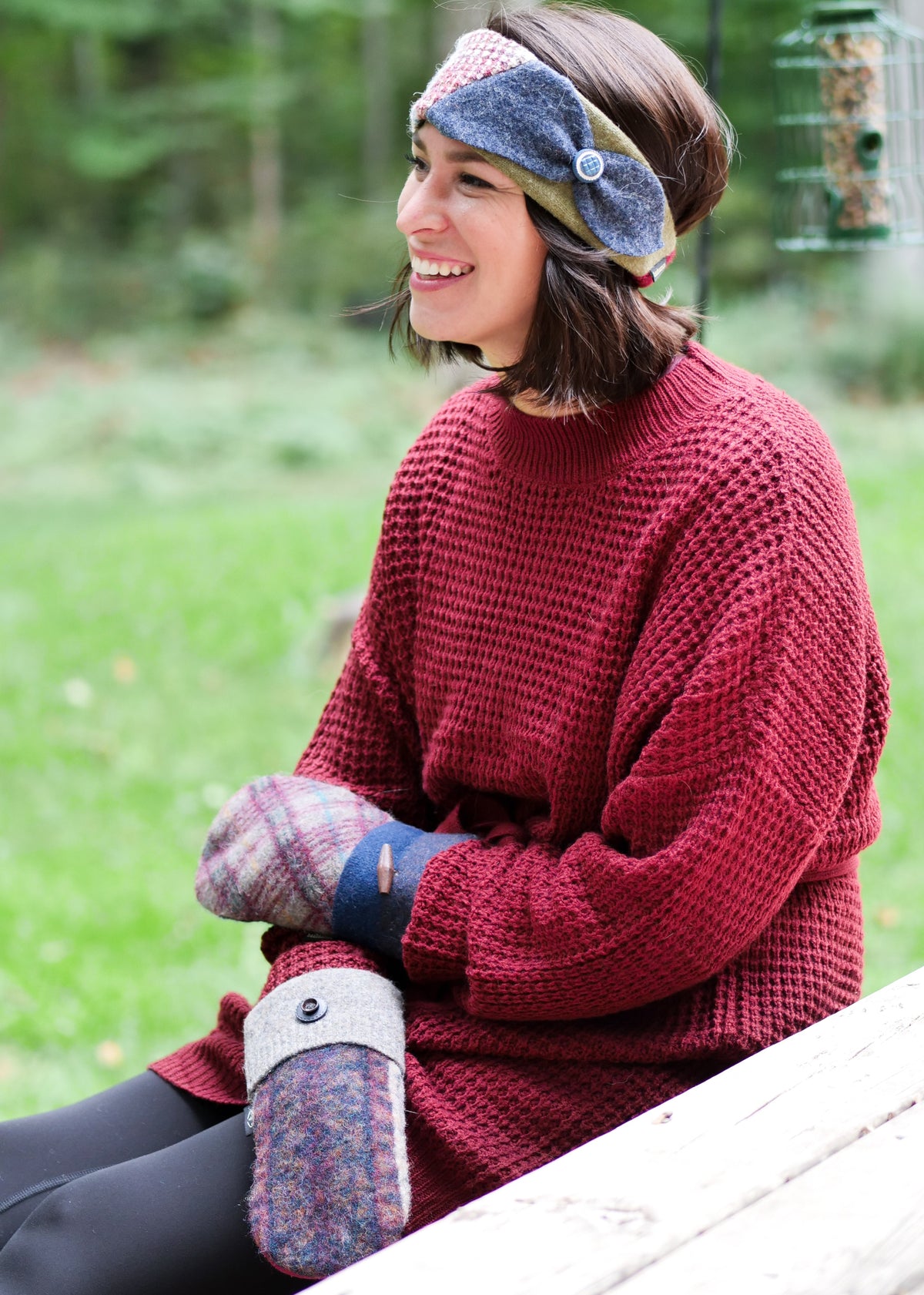A woman sitting outdoors on a wooden bench wearing a chunky red knit sweater, a blue and green patterned headband, sweater mittens, and black leggings, with a blurred green forest background.
