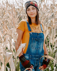 A woman standing in a cornfield wearing blue denim overalls over a mustard yellow graphic t-shirt, a patterned sweater headband, and upcycled wool fingerless gloves.