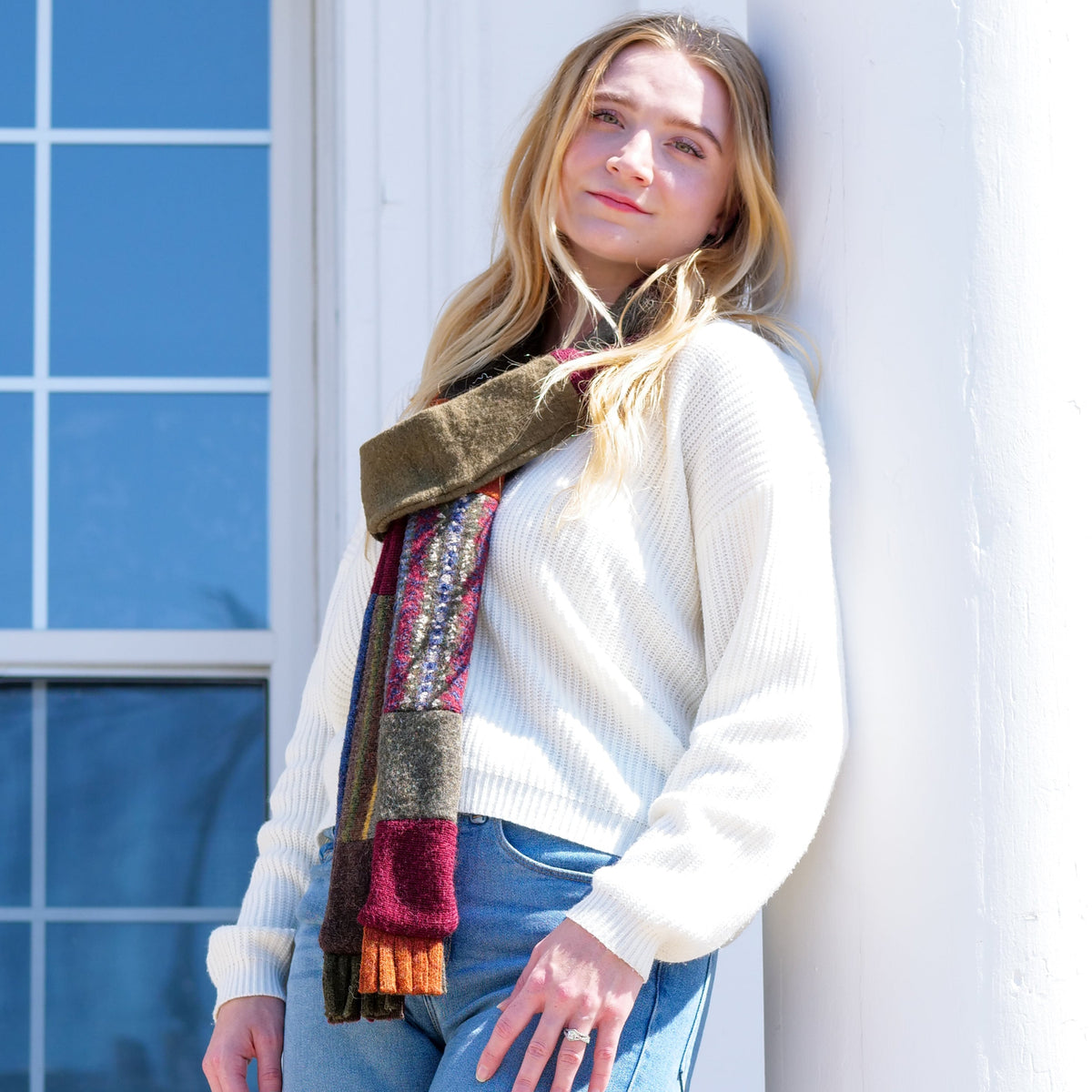 A woman wearing a cream ribbed knit sweater, blue jeans, and a multi-colored patchwork scarf leans against a white exterior wall next to a window.