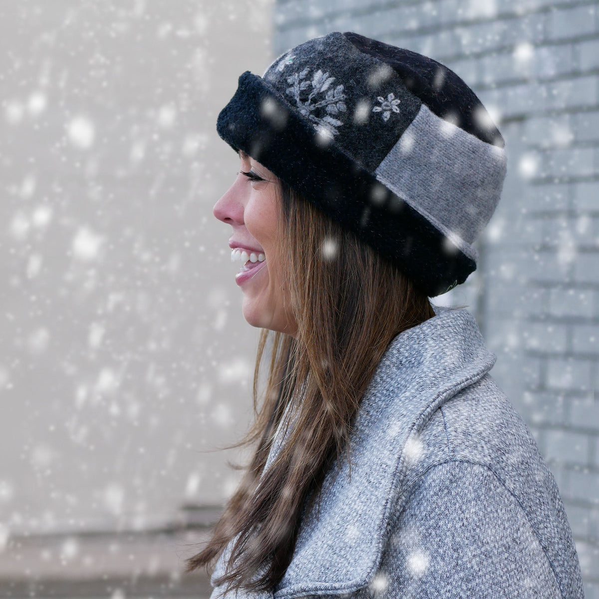 A woman in a gray textured coat wearing a black and gray pillbox hat with snowflake details, standing outdoors in falling snow.