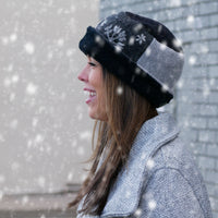 A woman in a gray textured coat wearing a black and gray pillbox hat with snowflake details, standing outdoors in falling snow.