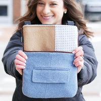 A woman holding up a baaabaazuzu compact zip bag with a patchwork design. The sleeve features a tan textured panel, a white and blue patterned panel, and a large light blue felt lower section with a front pocket.