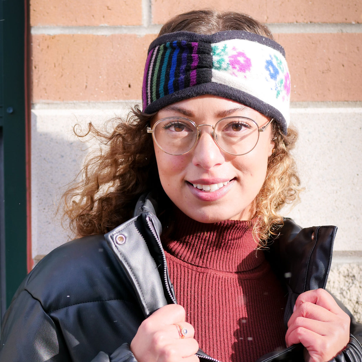 A woman wearing a black winter puffer jacket, a dark red ribbed turtleneck, and a colorful striped knit headband with a floral pattern, standing in front of a brick wall.