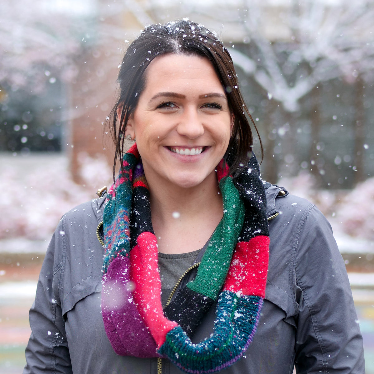 A smiling woman outdoors in falling snow, wearing a chunky multicolor sweater infinity scarf with green, red, pink, purple, and black sections, paired with a dark gray jacket.