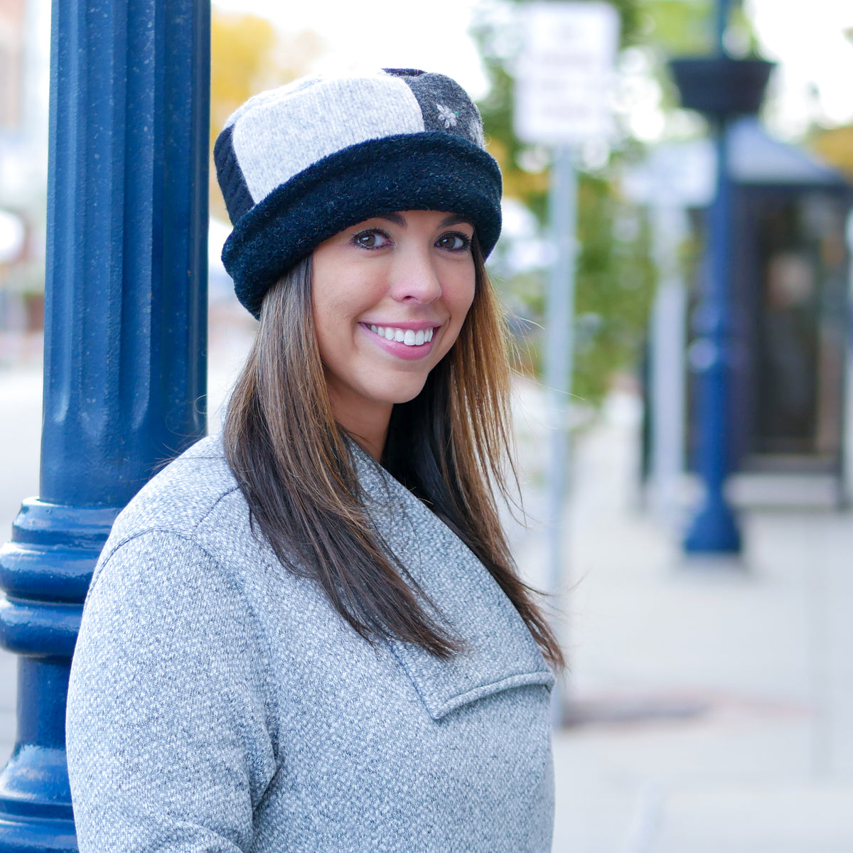 A smiling woman wearing a gray and black knit beanie hat and a gray textured coat, standing outdoors next to a blue pole with a blurred city street background.