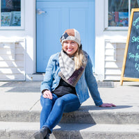 Sue Burns sits on concrete steps in front of a white storefront with a light blue door, wearing a beige bow hat, a patterned cowl scarf, a blue denim jacket, a black top, and blue jeans.