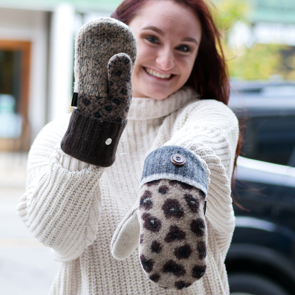 A smiling woman wearing a cream knit sweater holds up two patterned winter mittens. One mitten has a dark brown and black leopard print with a dark cuff, and the other has a beige and black leopard print with a blue-gray cuff and a button detail.