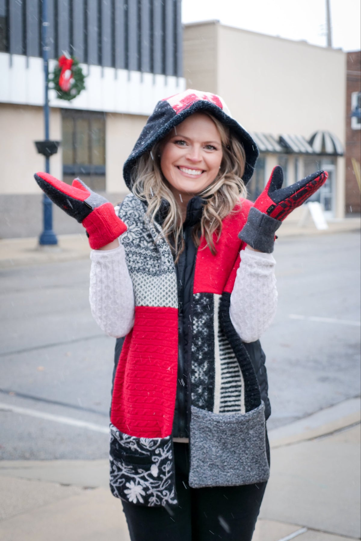 A woman smiling outdoors in a winter outfit featuring a red, black, and white hoodie scarf, coordinating knit mittens from baabaazuzu standing on a city street.