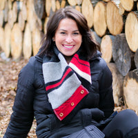 A smiling woman wearing a black puffer jacket and a thick wool scarf with red, gray, white, and black stripes sits outdoors in front of a stacked log wall.