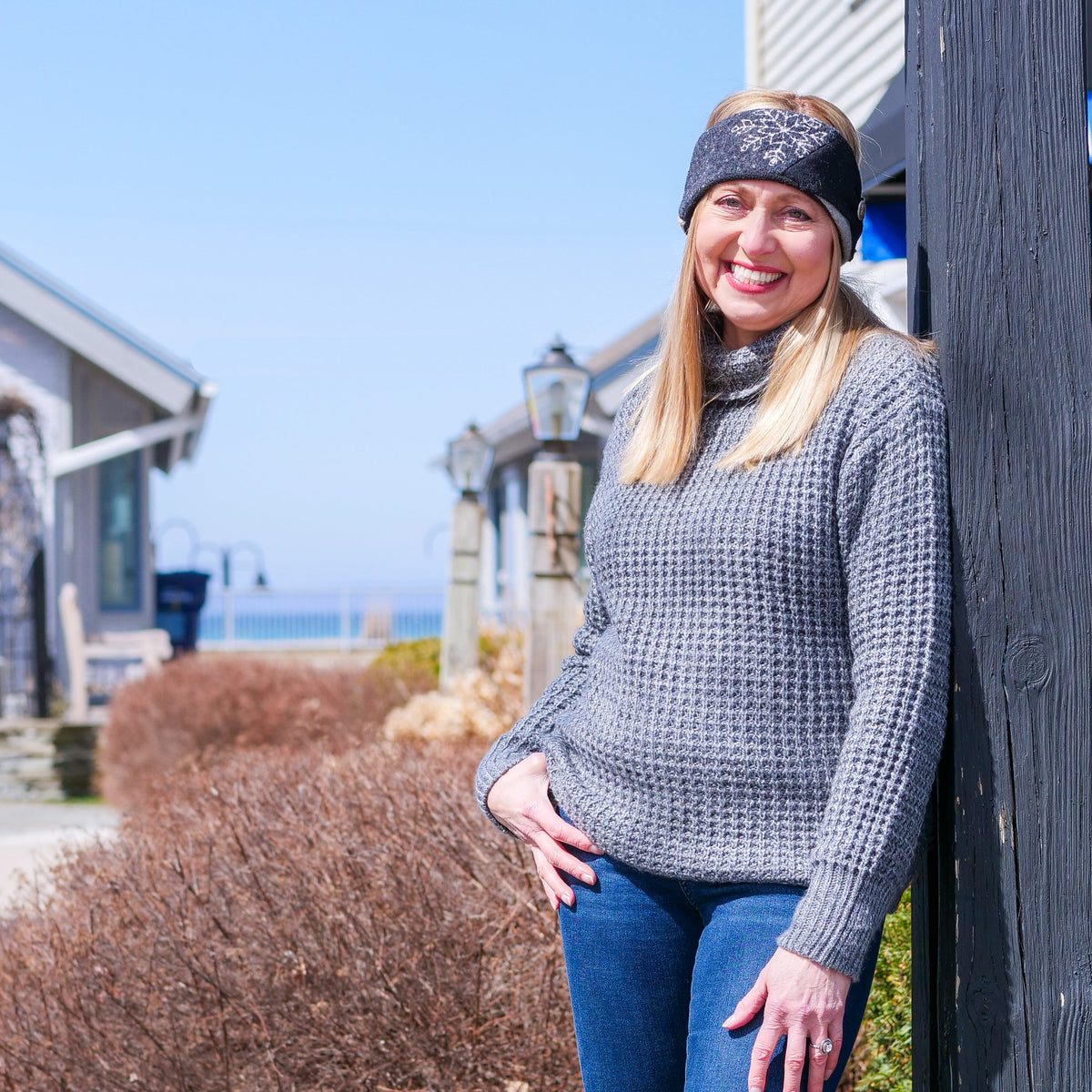 A woman wearing a gray waffle-knit sweater, blue jeans, and a black patterned headband stands outdoors leaning against a dark wooden post in a sunny coastal setting.