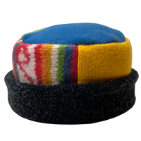 A handmade wool cap featuring a blue top panel, a yellow knit section, a multicolor striped patterned band, and a black fuzzy brim, shown against a plain white background.