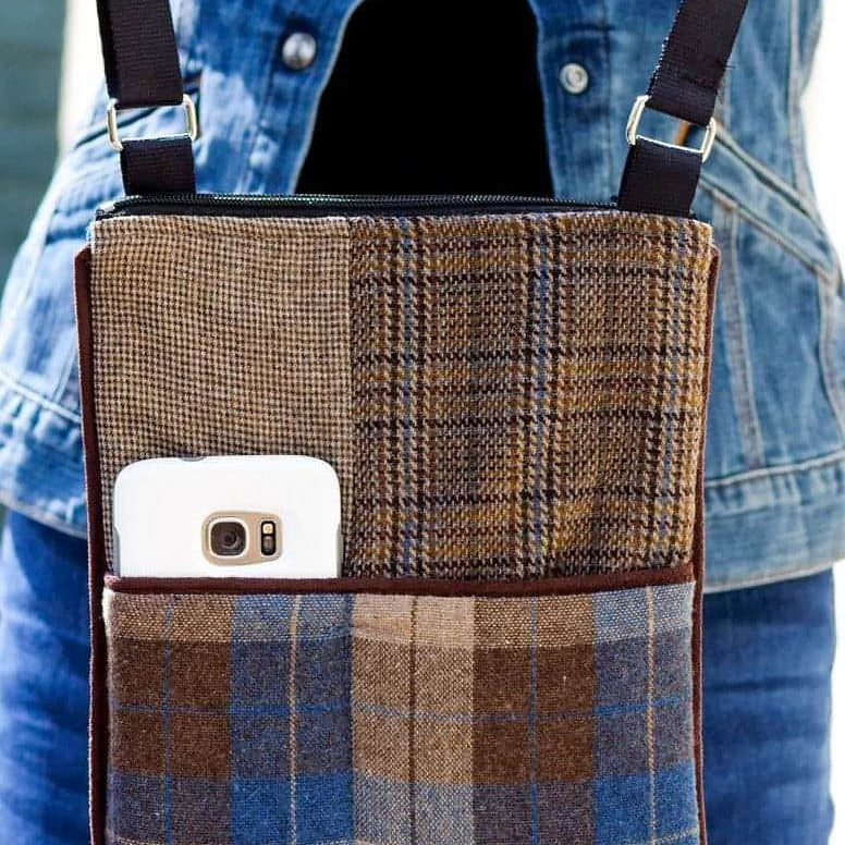 A close-up of a fabric crossbody phone bag with a patchwork design in brown, beige, and blue plaid. A white smartphone is shown in the front pocket, and a person wearing a blue denim jacket is visible in the background.